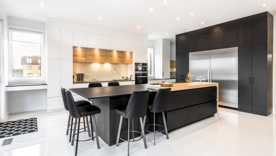 Cuisine moderne noire et blanche composée d'armoires en Thermoform accentuées de bois de chêne. L'îlot central est prolongé d'une table de manger autour de laquelle six tabourets sont disposés.