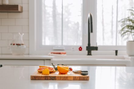 Plan cadré d'une cuisine Ateliers Jacob. Une planche à découper avec des mandarines est disposée sur le comptoir de l'îlot central. La cuisine est trés épurée et blanche.