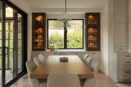 Salle à manger lumineuse avec table en bois clair, chaises texturées et vitrines rétroéclairées encadrant une grande fenêtre donnant sur la verdure.
