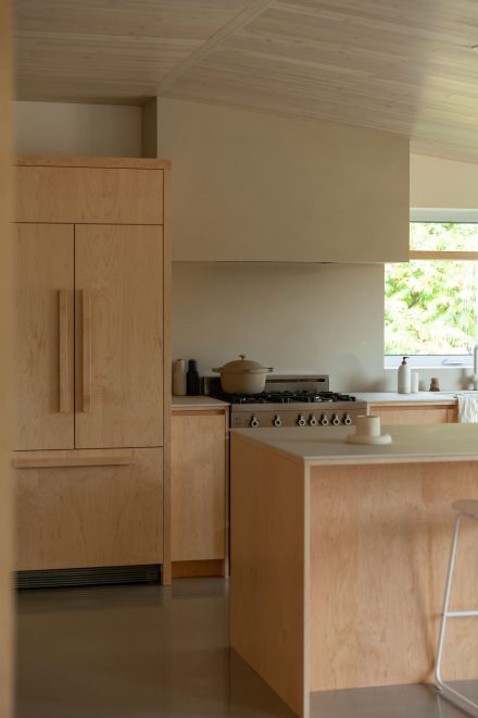 Cuisine lumineuse au design minimaliste, avec des armoires en bois pâle, un évier de style farmhouse blanc et une cuisinière à gaz, mise en valeur par une grande fenêtre offrant une vue sur la verdure extérieure.
