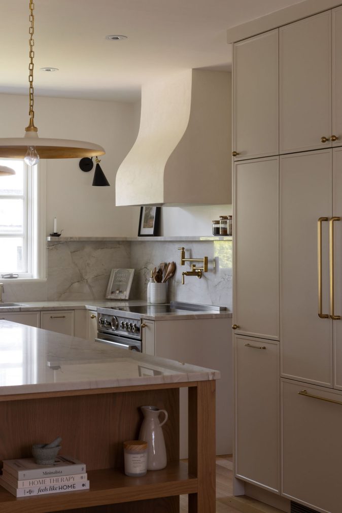 Cuisine haut de gamme conçue par Ateliers Jacob à Montréal, mariant des armoires crème avec des poignées en laiton, un îlot en bois clair et un comptoir en marbre. La hotte sculpturale et les luminaires suspendus ajoutent une touche artisanale et contemporaine à l’ensemble.