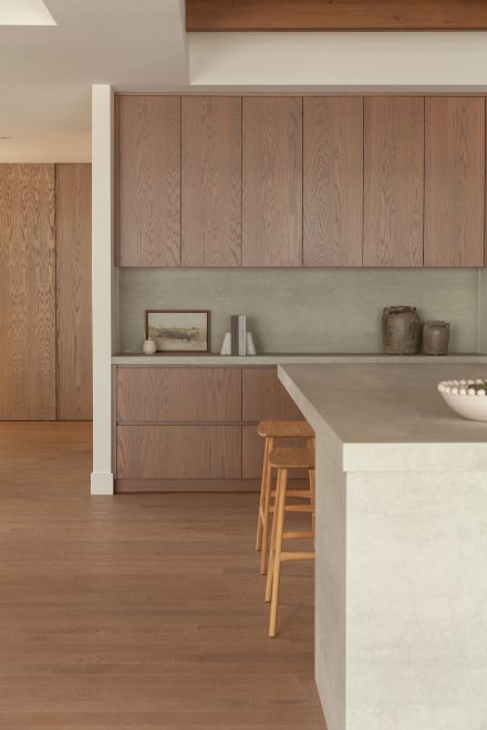Cuisine moderne haut de gamme signée Ateliers Jacob avec îlot en pierre claire, hotte dissimulée en béton, armoires pleine hauteur crème, placages de bois naturel et poutres apparentes au plafond, design minimaliste et chaleureux.