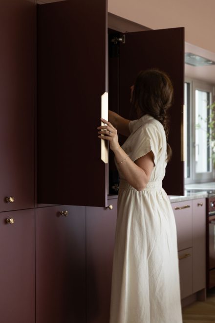 Femme ouvrant une armoire bourgogne sur mesure dans une cuisine Ateliers Jacob, mettant en valeur la fonctionnalité du rangement intégré et le design haut de gamme aux finitions soignées