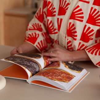 Une personne préparant des aliments dans une cuisine moderne et chaleureuse. L’espace met en valeur des matériaux naturels comme le bois clair et un design épuré. On aperçoit une cuisinière, un four de Our Place, et des accessoires de cuisine soigneusement sélectionnés, créant une atmosphère conviviale et fonctionnelle.