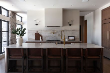 Cuisine sur mesure en chêne conçue par Ateliers Jacob au Québec, avec grand îlot central en pierre naturelle, robinetterie en laiton, hotte encastrée et dosseret en pierre, dans un aménagement lumineux aux lignes épurées.