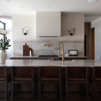 Cuisine sur mesure en chêne conçue par Ateliers Jacob au Québec, avec grand îlot central en pierre naturelle, robinetterie en laiton, hotte encastrée et dosseret en pierre, dans un aménagement lumineux aux lignes épurées.