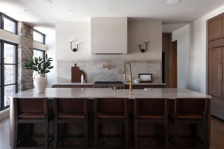 Cuisine sur mesure en chêne conçue par Ateliers Jacob au Québec, avec grand îlot central en pierre naturelle, robinetterie en laiton, hotte encastrée et dosseret en pierre, dans un aménagement lumineux aux lignes épurées.