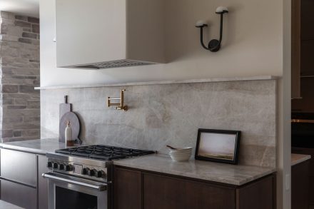 Cuisine sur mesure en chêne conçue par Ateliers Jacob au Québec, avec grand îlot central en pierre naturelle, robinetterie en laiton, hotte encastrée et dosseret en pierre, dans un aménagement lumineux aux lignes épurées.