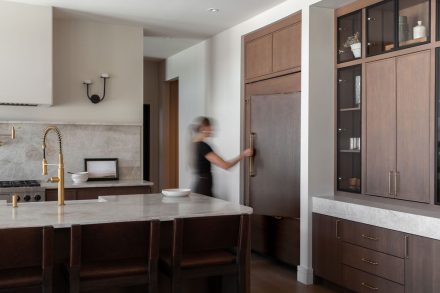 Cuisine sur mesure en chêne conçue par Ateliers Jacob au Québec, avec grand îlot central en pierre naturelle, robinetterie en laiton, hotte encastrée et dosseret en pierre, dans un aménagement lumineux aux lignes épurées.