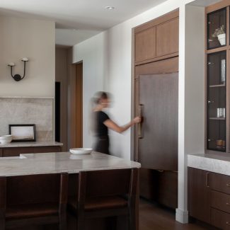 Cuisine sur mesure avec îlot en pierre naturelle, armoires en bois foncé, vitrines encadrées de verre et réfrigérateur intégré dissimulé derrière des panneaux assortis.