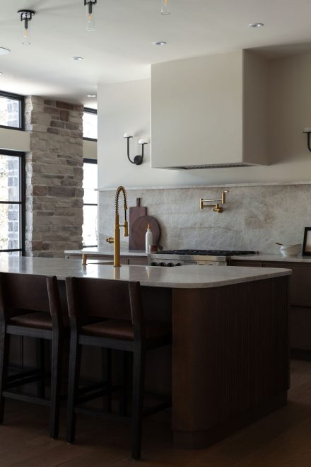 Cuisine sur mesure en chêne conçue par Ateliers Jacob au Québec, avec grand îlot central en pierre naturelle, robinetterie en laiton, hotte encastrée et dosseret en pierre, dans un aménagement lumineux aux lignes épurées.