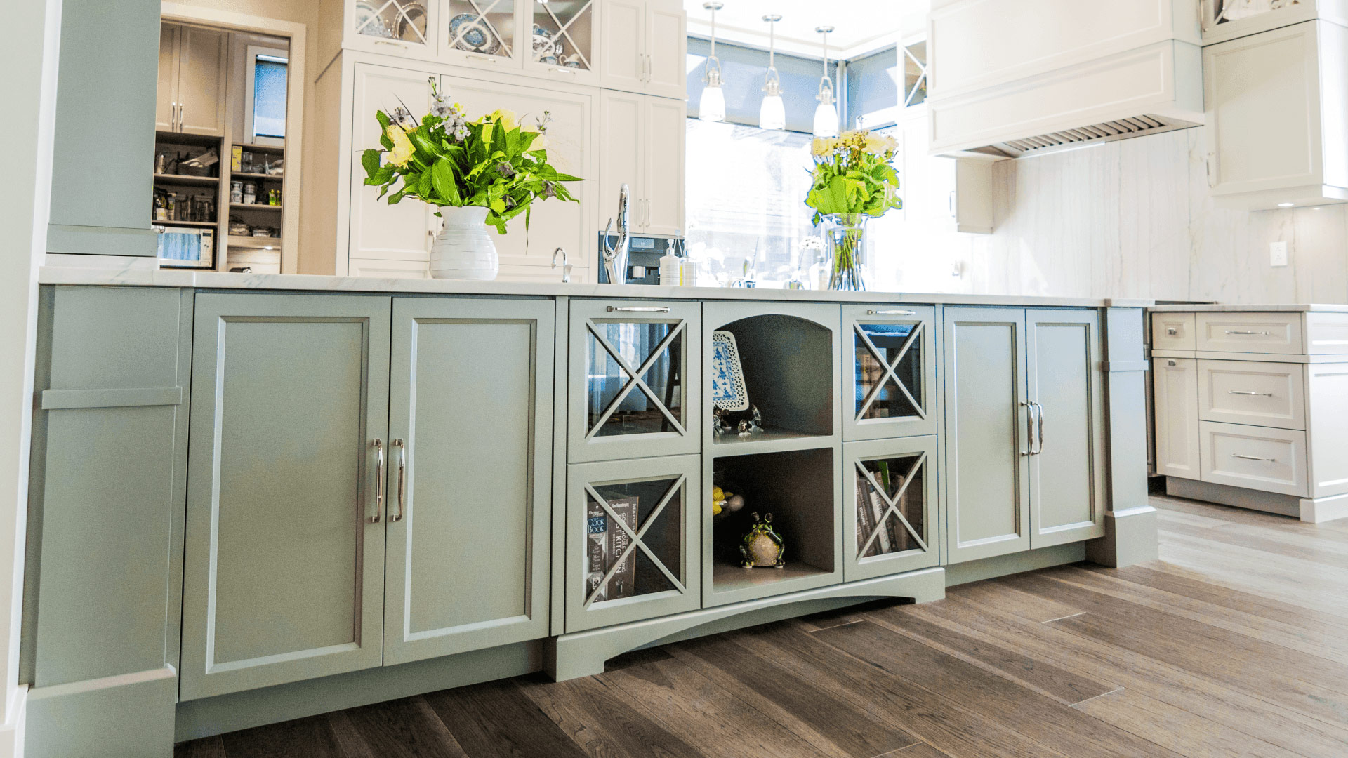 Kitchen cabinets of the central island of a kitchen in soft green color. Some of the wardrobes feature glass doors.