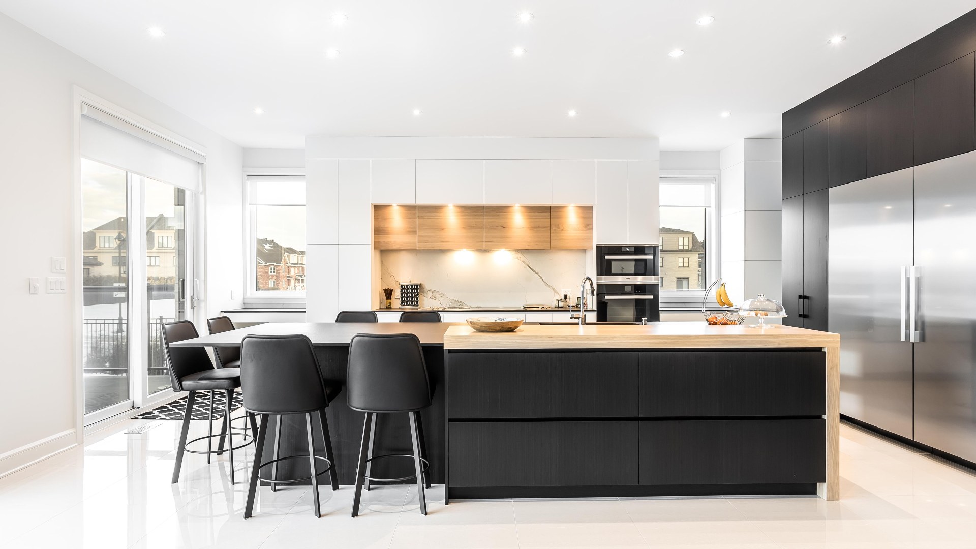 Cuisine moderne noire et blanche composée d'armoires en Thermoform accentuées de bois de chêne. Des luminaires sont iintégrés aux meubles de cuisines pour éclairer les plans de travail et comptoirs directement.